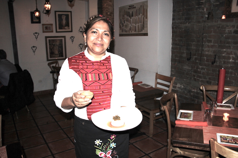 La Chef Olga Cabrera muy emocionada de presentar por primera vez en Caf&eacute; Frida de Nueva York, la aut&eacute;ntica cocina mixteca, sostiene un Nicuatole con nopalitos en alm&iacute;bar y una peque&ntilde;a j&iacute;cara con whisky de ma&iacute;z Pierde Almas Ancestral.
