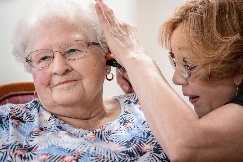 Janie York examina el o&iacute;do de Elaine Martin en SilverRidge Assisted Living, en Gretna, Nebraska, en agosto. Martin ten&iacute;a bastante cera en sus o&iacute;dos, antes que York le realizara una limpieza y comenzara a usar aud&iacute;fonos. Ahora, York le limpia los o&iacute;dos a Martin con regularidad. (Chris Machian para KHN)