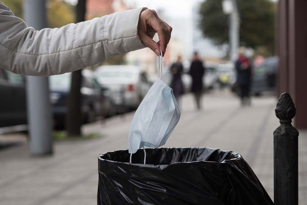 A woman throws a facemask at a public trash can on a city street mascarilla