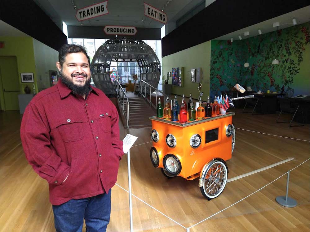 MONXO MUESTRA COMIDA 1 Monxo Lopez, junto a la obra del artista puertorriqueño Miguel Luciano, que conmemora la innovación de los vendedores ambulantes latinos, transformando un carrito de venta de hielo raspado, -conocido en el Caribe como piragua-, en un triciclo con sistema de sonido, pantallas y luces.