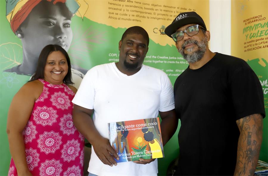 Miguel Quiñones (c) posa con el libro El vejigante come coco, junto a su autora, la activista Maricruz Rivera Clemente (i), y el artista plástico Celso González