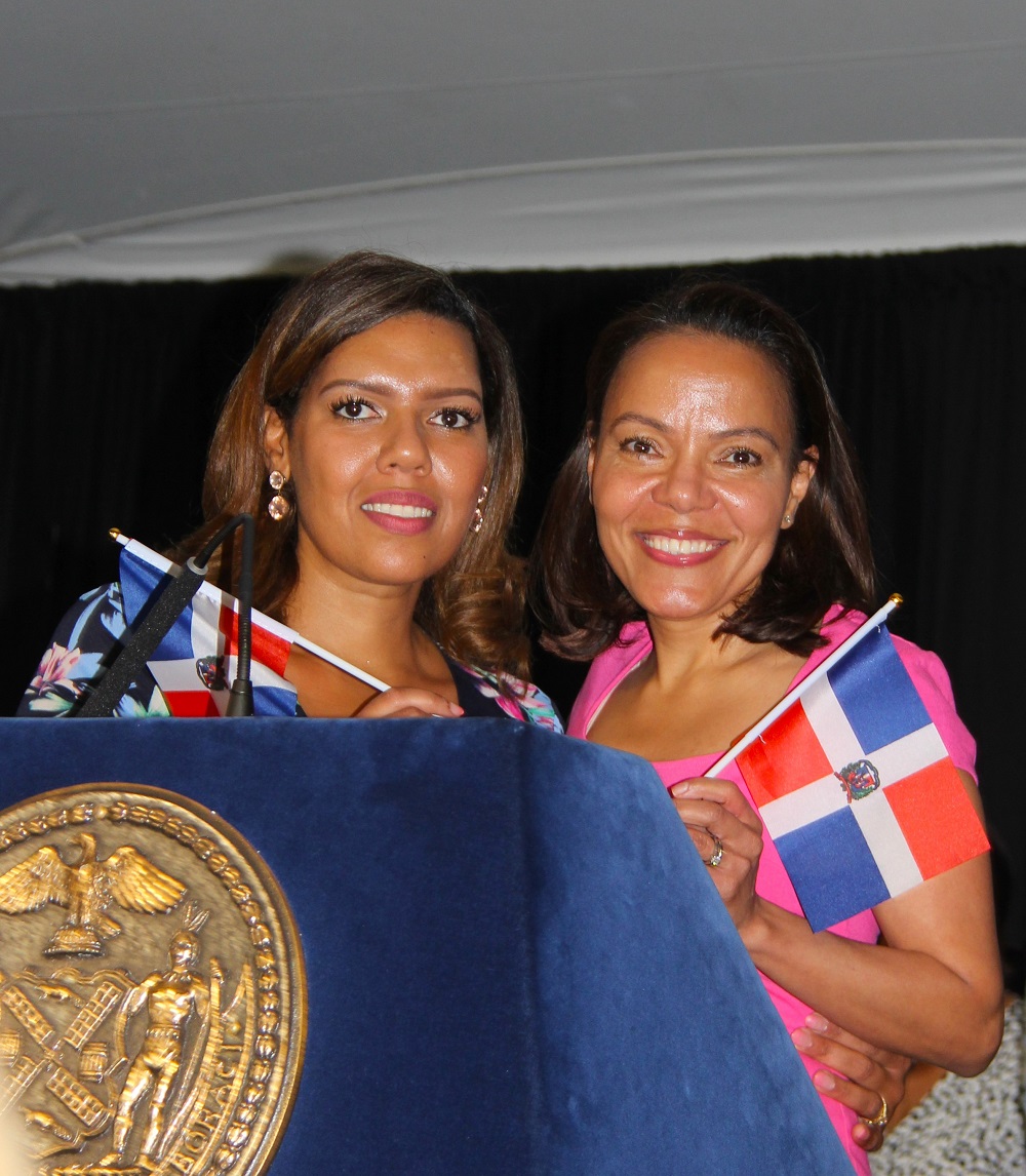 Ana Almanzar, la primera dominicana de la historia de la ciudad en ser nombrada vicealcaldesa de Iniciativas Estratégicas por Eric Adams, a la derecha de la gráfica, junto a la efusiva maestra de ceremonias, orgullosas portan la bandera dominicana que ha sido la reina de los festejos de agosto.
