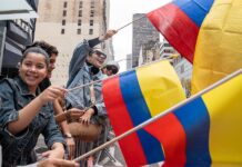 Varias personas ondean banderas colombianas mientras observan el desfile del Día de la Hispanidad