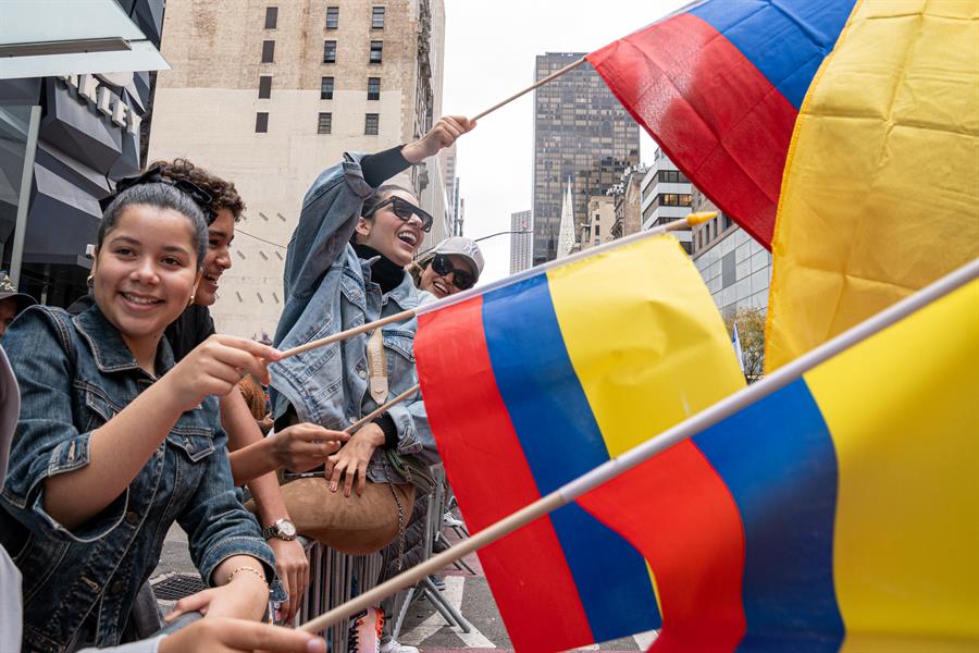 Varias personas ondean banderas colombianas mientras observan el desfile del D&iacute;a de la Hispanidad