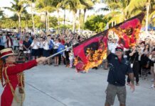 Jai Somers (i) sosteniendo una bandera de advertencia de huracán incendiada y Paul Menta (d) rociándola con ron